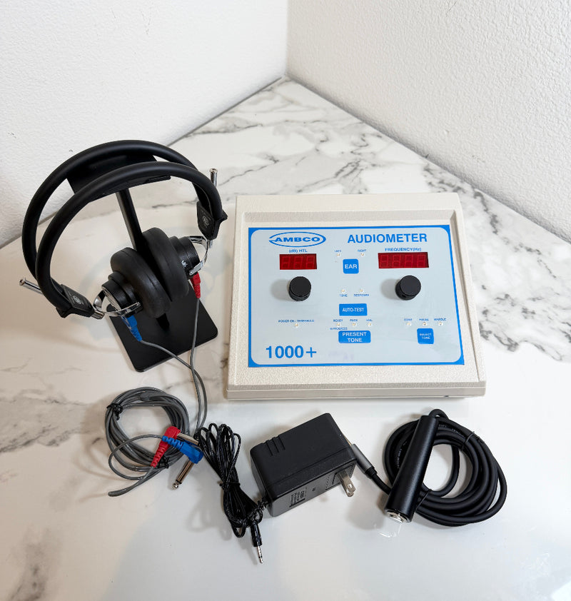 An AMBCO 1000+ audiometer testing device with a beige control panel, black headphones on a stand, cables, and a power adapter, arranged on a white marble-patterned surface against a white wall corner.