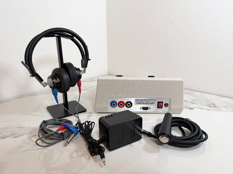 A portable audiometer device set on a white marble-like surface against a white wall. The set includes a beige rectangular electronic unit with red, yellow, blue, and white audio jacks, black headphones on a stand, a black power adapter, and a black wired controller.