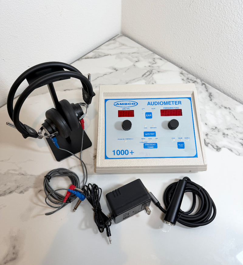 An AMBOO 1000+ audiometer device with its attached headphones, cables, and power adapter, arranged on a white marble-patterned surface against a white wall corner.