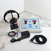 A AMBCO 1000+ audiometer device with a beige control box, blue front panel, black headphones on a stand, black cables, and a black power adapter arranged on a white marble-patterned surface against a white wall.