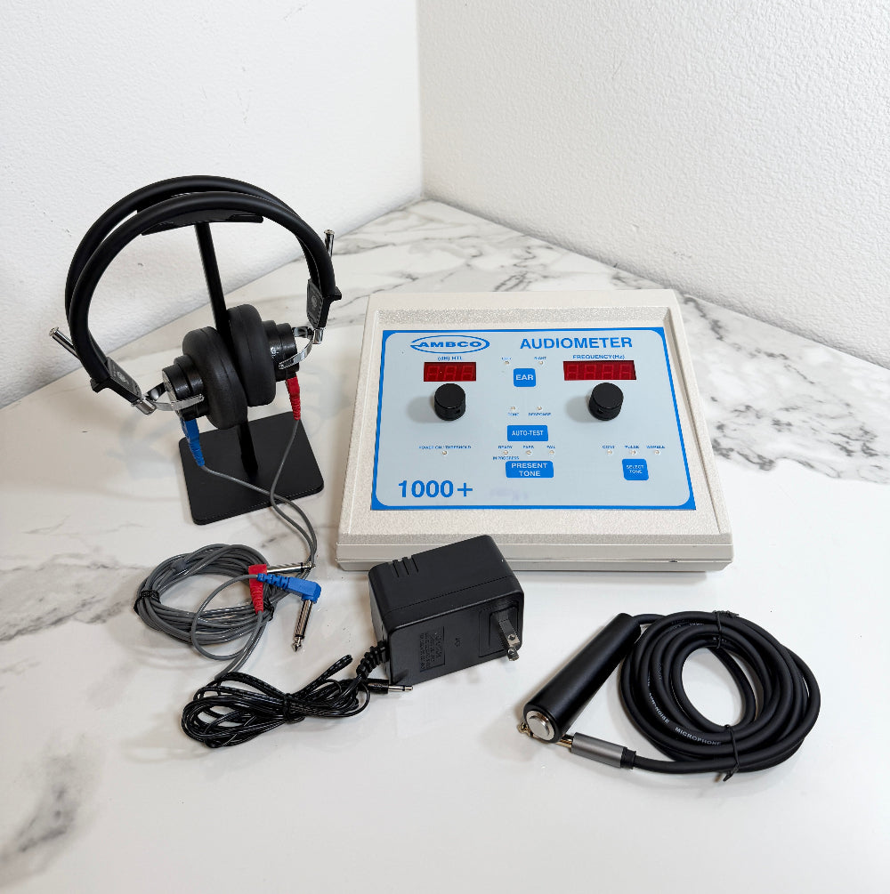 A AMBCO 1000+ audiometer device with a beige control box, blue front panel, black headphones on a stand, black cables, and a black power adapter arranged on a white marble-patterned surface against a white wall.