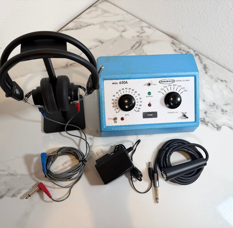 An AMBCO 650A Audiometer device with a blue metal housing and white faceplate, shown with a black headphone stand, headphones, power adapter, and audio cables on a white marble surface.