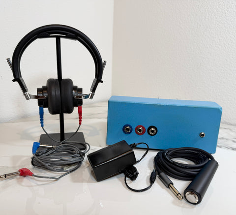 A pair of black over-ear headphones displayed on a stand, shown with a blue audio interface box, power adapter, and cables on a white surface against a white wall.