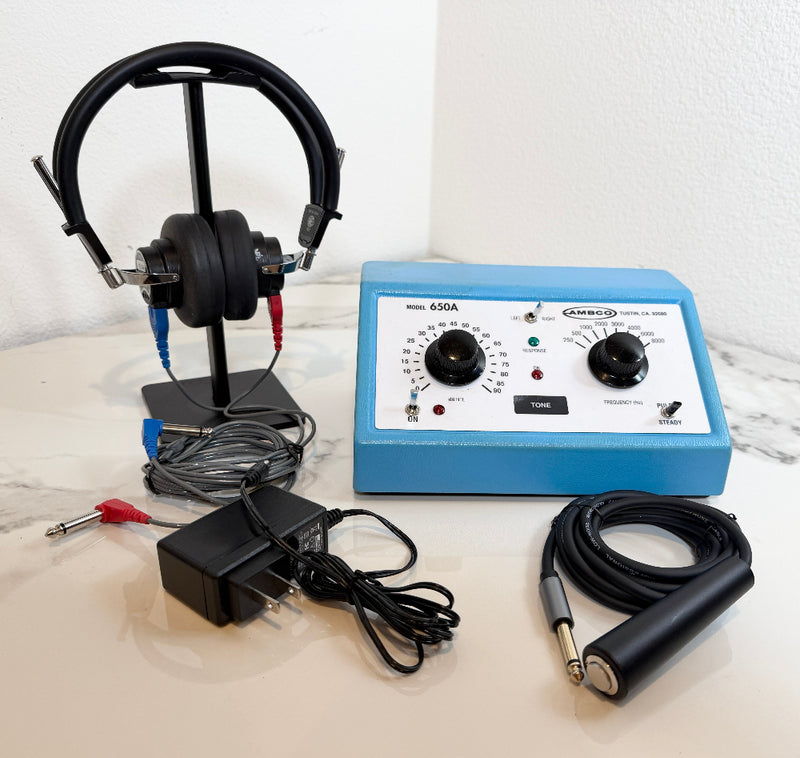 An AMBCO 650A Audiometer with a blue control box, black headphones on a stand, a black power adapter, and a black cable with a silver and black electrode, arranged on a light marble surface against a white wall.
