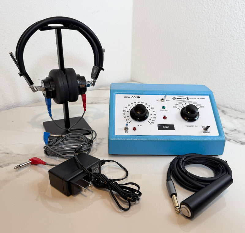 An AMBCO 650A Audiometer  kit with a blue control box, black headphones on a stand, an adjustable cable, a power adapter, and a handheld electrode pad, displayed on a light marble surface against a white wall.
