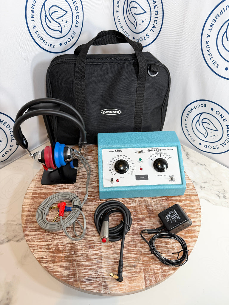 AMCO audiometer  kit with a light blue control unit, black headphones with red and blue ear cups, cables, an adapter, and a black carrying bag, arranged on a round wooden tray in front of a branded white backdrop.