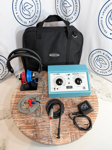 AMCO audiometer  kit with a light blue control unit, black headphones with red and blue ear cups, cables, an adapter, and a black carrying bag, arranged on a round wooden tray in front of a branded white backdrop.