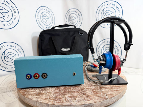 AMBCO audiometer device with three audio jacks on the front, displayed alongside a black carrying case, a black headphone stand with blue and red over-ear headphones, and a round wooden pedestal against a branded white backdrop.