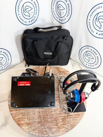 AMBCO audiometer setup displayed on a round wooden table, including a black monitor base with a red label, a pair of black and blue headphones on a stand, and a black carrying case.