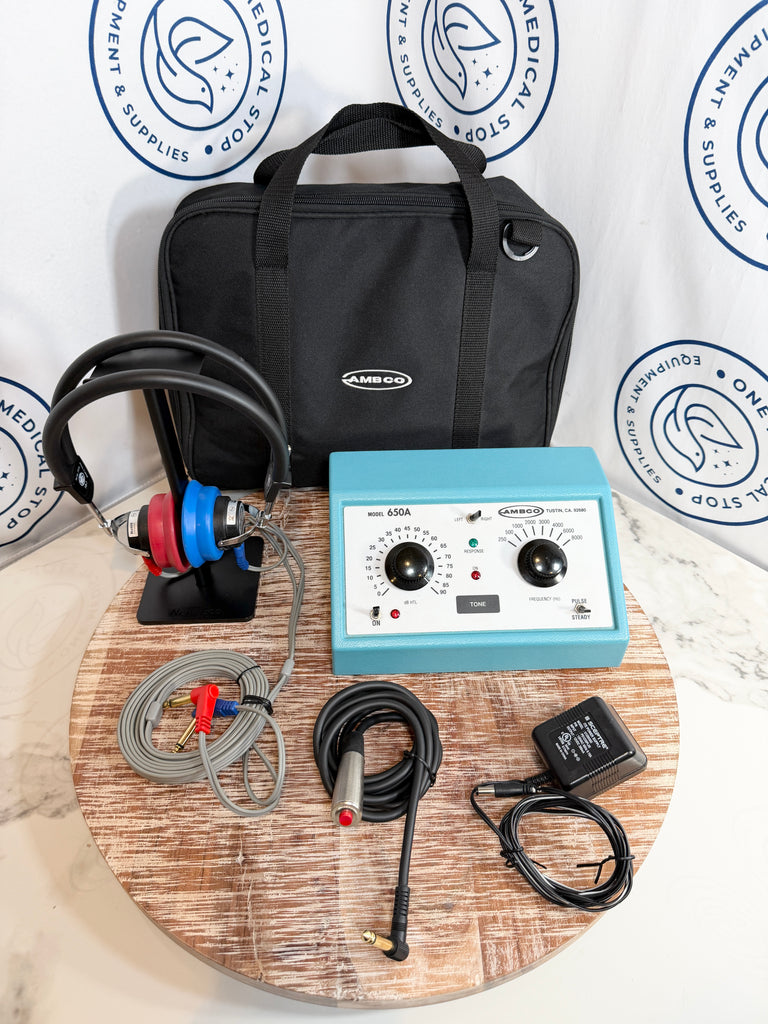 AMBCO audiometer kit with a light blue sound box, black headphones with red and blue ear cups, a black carrying case, cables, and an external power adapter, arranged on a round wooden board in front of a branded backdrop.