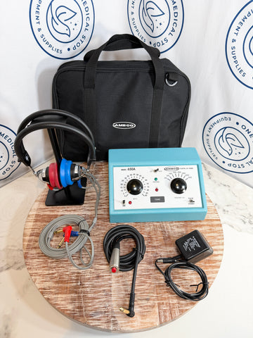 AMBCO audiometer kit with a light blue sound box, black headphones with red and blue ear cups, a black carrying case, cables, and an external power adapter, arranged on a round wooden board in front of a branded backdrop.