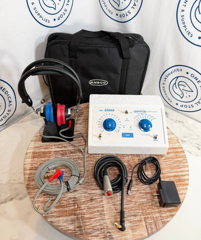 An AMBCO audiology equipment kit including a white sound generator console with blue knobs, a pair of red and blue headphones on a stand, a black carrying case, a gray cable, a metal probe, and an external power adapter, arranged on a round wooden table in front of a branded backdrop.