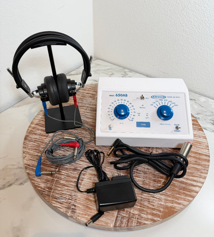A white and black TENS unit muscle stimulator with attached headphones, cables, and an external power adapter, arranged on a round wooden board against a white wall.