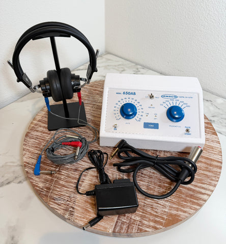 An AMBCO 650AB Audiometer with an attached black over-ear headphone, gray electrode leads, power adapter, and power cord, arranged on a round wooden tray against a white marble-patterned surface and white wall.