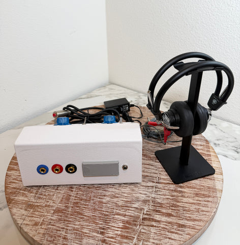 A white rectangular AMBCO 650AB Audiometer device with blue, red, and yellow audio jacks sits on a round wooden tray next to a black over-ear monitor on a stand, with cables and an adapter in the background.