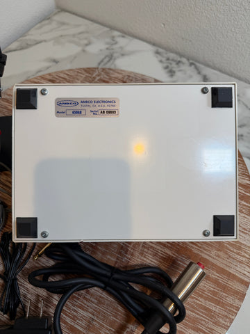 An AMBCO 650AB Audiometer power supply unit shown from the back, featuring a white metal casing with black corner protectors and a small illuminated yellow indicator light. It is resting on a wooden surface with its power cables and connectors visible.