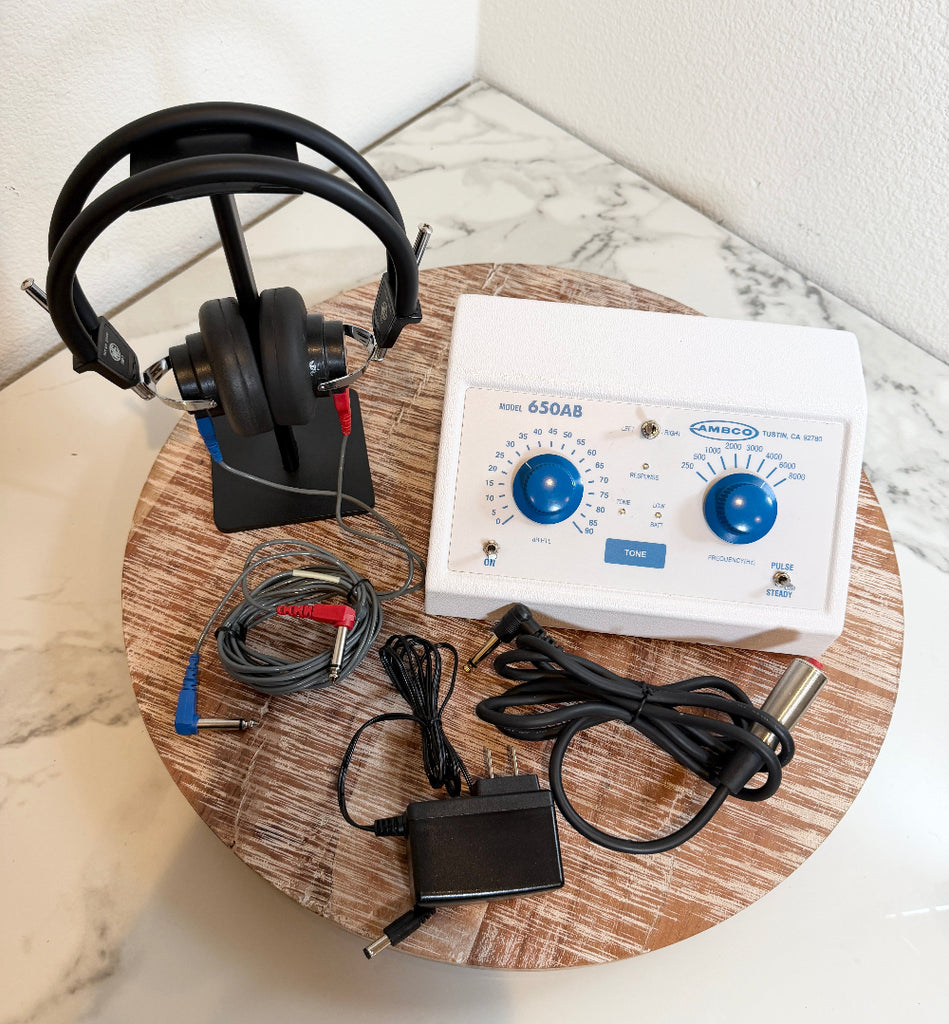 An AMBCO 650AB Audiometer device with attached black headphones on a stand, cables, and an adapter, arranged on a round wooden tray against a white marble-patterned surface and wall.
