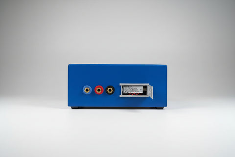 A blue audiometer with three colored RCA-style jacks and a small battery compartment with its door open, placed on a wooden surface against a light wall.