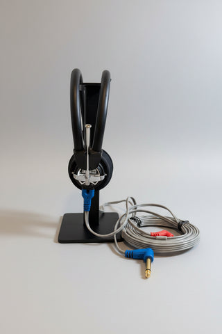 A black headset on a matching display stand, with a coiled gray cable and blue audio adapter on a wooden tabletop against a light wall.