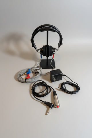 A pair of black over-ear headphones on a display stand, shown with attached gray wiring, red and blue connector clips, a black power adapter, and a silver-and-black audio cable on a round wooden board against a light wall.