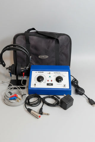 A AMBCO audiometer displayed on a round wooden board including black cables, a metal probe, a black carrying bag, and a headset.