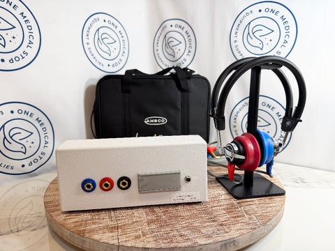 An AMBCO audiometer displayed on a round wooden table, featuring a white control unit with red, blue, and yellow connectors, a black AMBCO carrying case, a headphone on a stand. 