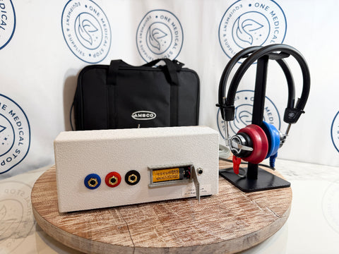 An AMBCO audiometer featuring blue, red, and black audio jacks and a yellow display window, displayed on a wooden stand next to a black carrying case and over-ear headphones on a stand.