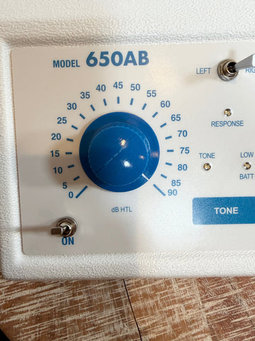 A close-up of the AMBCO audiometer control panel, model 650AB, featuring a large blue rotary volume knob with dB HTL markings, toggle switches, and a blue tone button on a white faceplate.