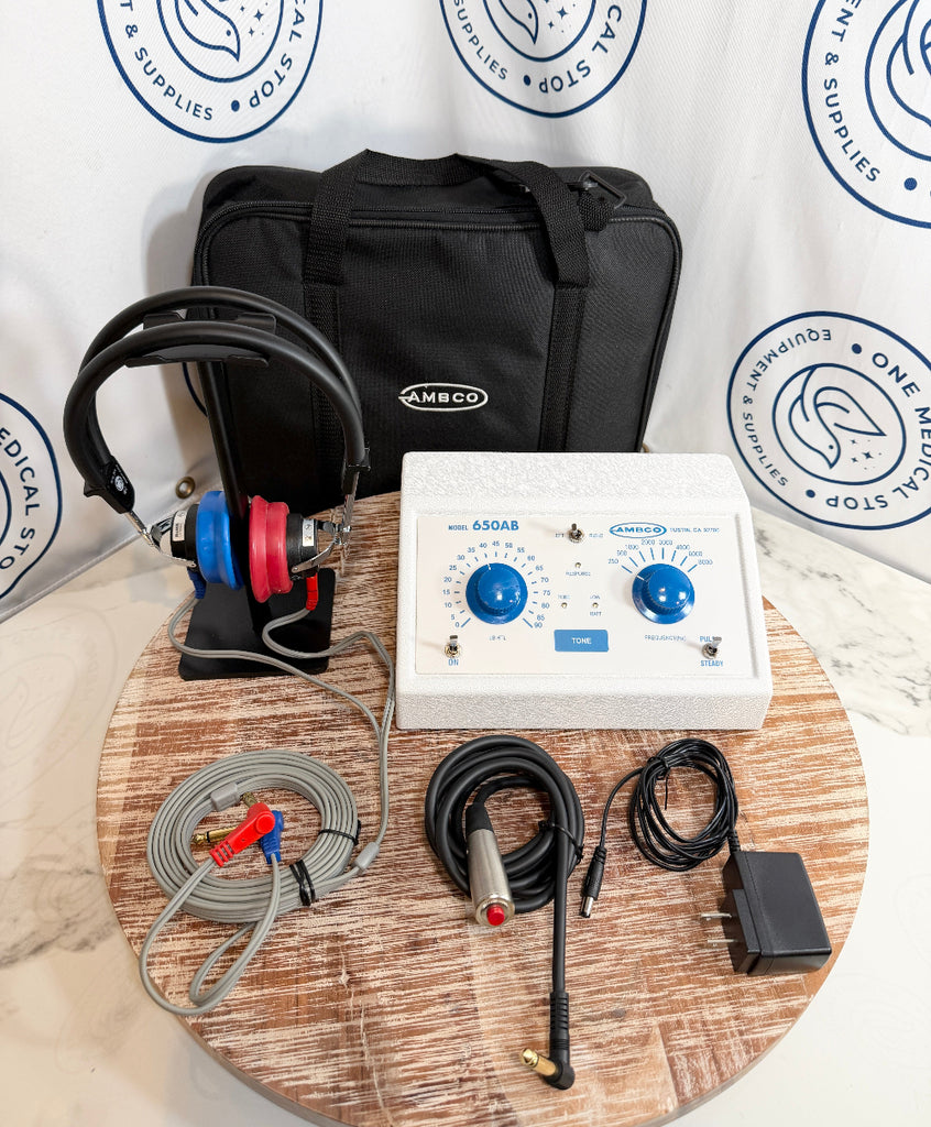 Ambco 650AB EKG/ECG monitor kit displayed on a round wooden tray, including a white control unit with blue dials, a black carrying case, a black and red electrode lead, gray cables, a metal probe, and an adapter, set against a branded white backdrop.