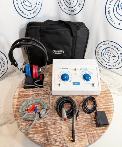 Ambco 650AB EKG/ECG monitor kit displayed on a round wooden tray, including a white control unit with blue dials, a black carrying case, a black and red electrode lead, gray cables, a metal probe, and an adapter, set against a branded white backdrop.