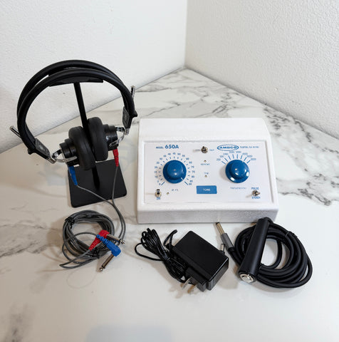 Ambco 650A Portable Screening Audiometer with a white control panel, blue dials, attached black headphones on a stand, electrode leads, a power adapter, and a TENS pad holder, arranged on a white marble-patterned surface against a white wall.