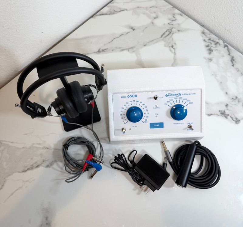 Ambco 650A Portable Screening Audiometer with attached black over-ear headphones, electrode leads, power adapter, and coiled cable, arranged on a white marble-patterned surface.