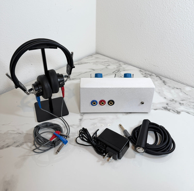 Ambco 650A Portable Screening Audiometer testing kit displayed on a white marble-patterned surface against a white wall. The kit includes a white rectangular electronic device with red, blue, and black audio jacks, a pair of black over-ear headphones on a stand, a black power adapter, a black cable with a silver plug, and several small clear plastic rings.