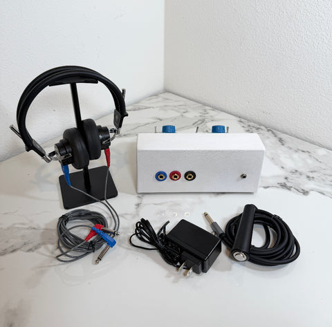 Ambco 650A Portable Screening Audiometer testing kit displayed on a white marble-patterned surface against a white wall. The kit includes a white rectangular electronic device with red, blue, and black audio jacks, a pair of black over-ear headphones on a stand, a black power adapter, a black cable with a silver plug, and several small clear plastic rings.