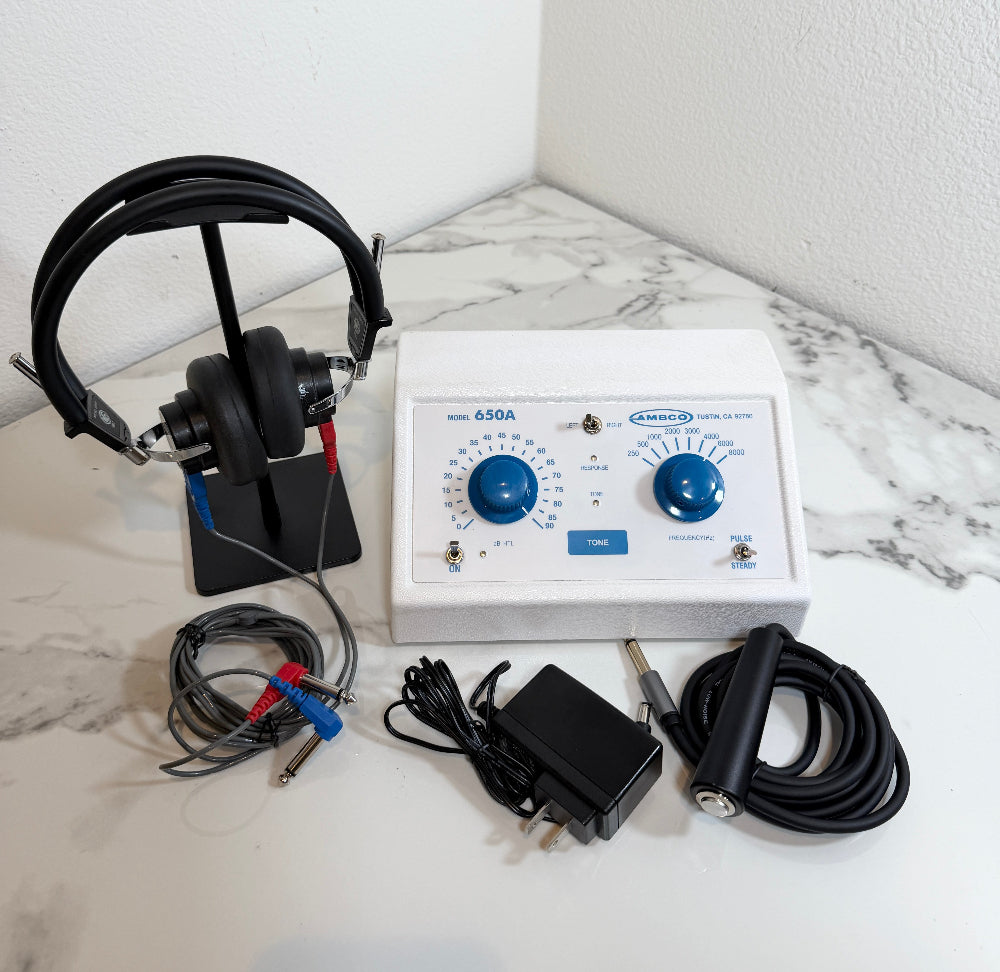 Ambco 650A Portable Screening Audiometer with a white control panel, blue dials, attached black cables, a pair of black over-ear headphones on a stand, and a black power adapter, arranged on a white marble-patterned surface against a white wall.