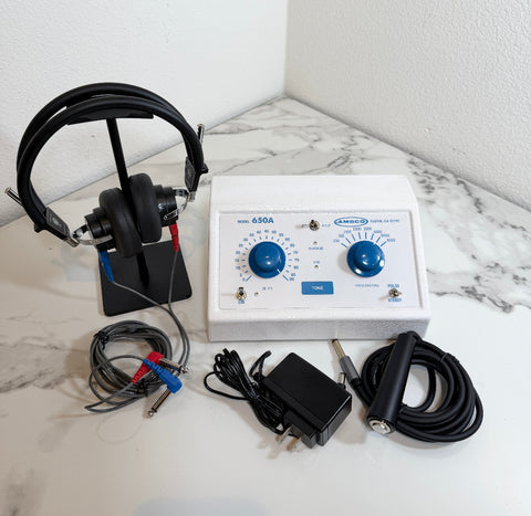 Ambco 650A Portable Screening Audiometer with a white control panel, blue dials, attached black cables, a pair of black over-ear headphones on a stand, and a black power adapter, arranged on a white marble-patterned surface against a white wall.