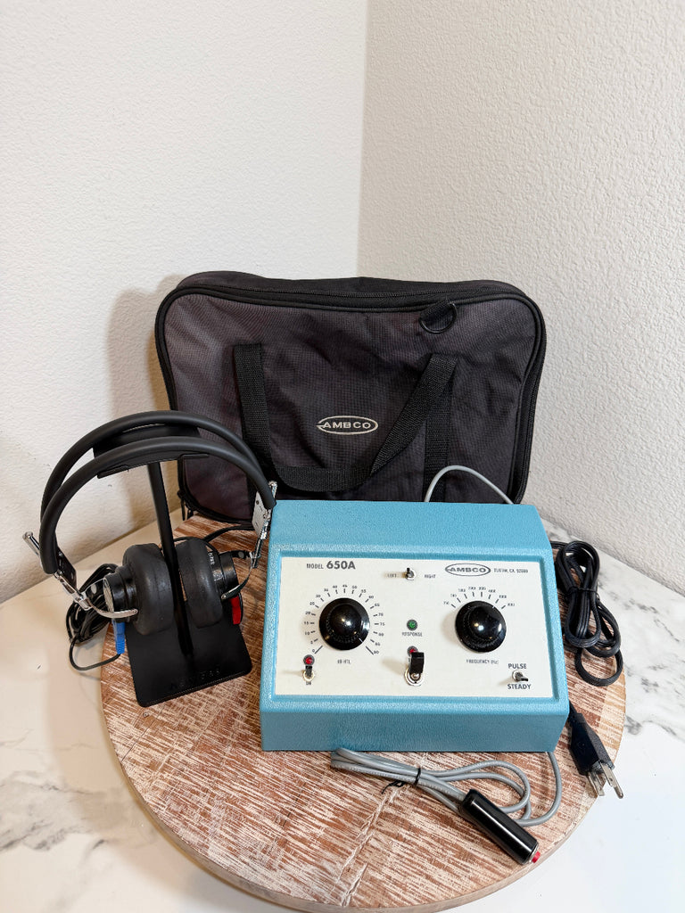 A blue and white AMBCO 650A audiometer device displayed on a round wooden tray, accompanied by black headphones, a black carrying case, and cables, set against a white wall corner.