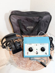 A MBCO 650A audiometer device with a light blue body, black dials, and attached headphones, displayed on a round wooden tray with its black carrying case behind it.