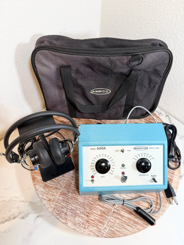 A MBCO 650A audiometer device with a light blue body, black dials, and attached headphones, displayed on a round wooden tray with its black carrying case behind it.