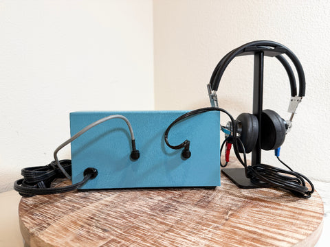 A light blue rectangular MBCO 650A audiometer device with attached black cables and a pair of black over-ear headphones on a black stand, displayed on a round wooden surface against a plain off-white wall.