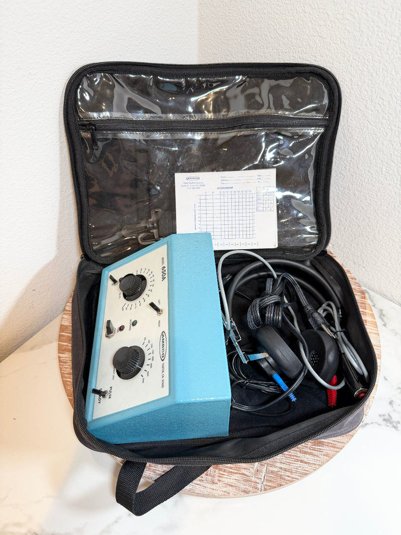 A blue and white AMBCO 650A audiometer device inside an open black zippered carrying case, shown with attached black wires and a paper chart on a round wooden tray against a white wall.
