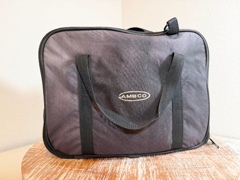A black AMBCO soft-sided carrying bag with black handles and trim, resting on a wooden surface against a plain light wall.