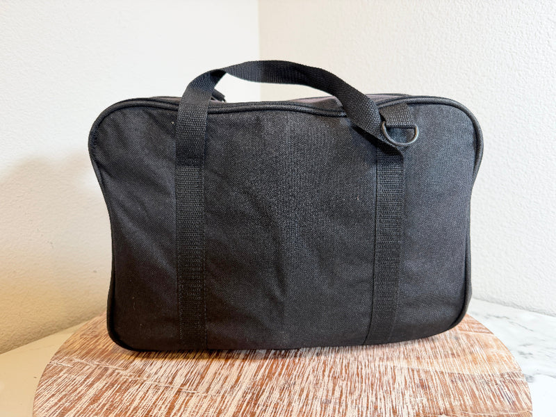 A black AMBCO soft-sided carrying bag with black handles and trim, resting on a wooden surface against a plain light wall. 