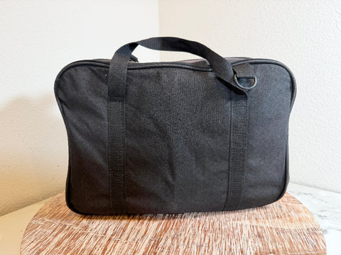 A black AMBCO soft-sided carrying bag with black handles and trim, resting on a wooden surface against a plain light wall. 