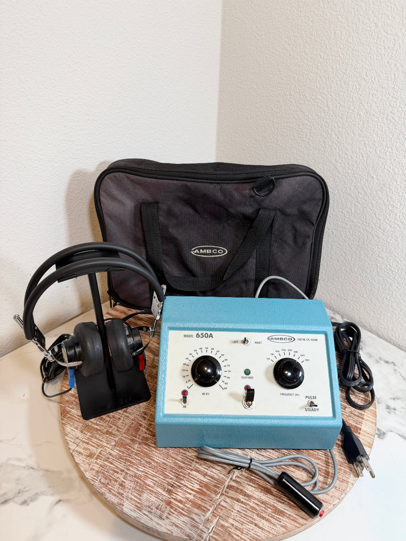 A blue and white Ambro 650A audiometer device with attached black headphones, shown with a black carrying case and cords on a round wooden tray against a white wall.