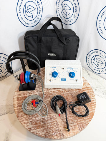 AMCO audiometer kit arranged on a round wooden tray, including a white control unit with blue knobs, black headphones with red and blue ear cups, gray cable leads, black electrode probes, a black power adapter, and a black carrying bag. 