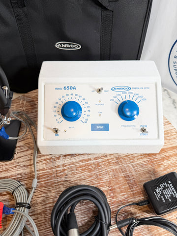 An AMBCO Model 650A audiometer with two blue rotary dials on a white front panel, shown on a wooden surface with cables, an adapter, and a black AMBCO carrying bag in the background.