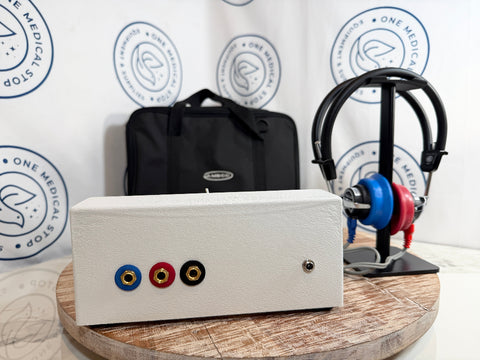AMBCO audiometer with three colored audio jacks on the front, displayed on a round wooden stand. Behind it is a black carrying case, and to the right is a pair of headphones on a stand.