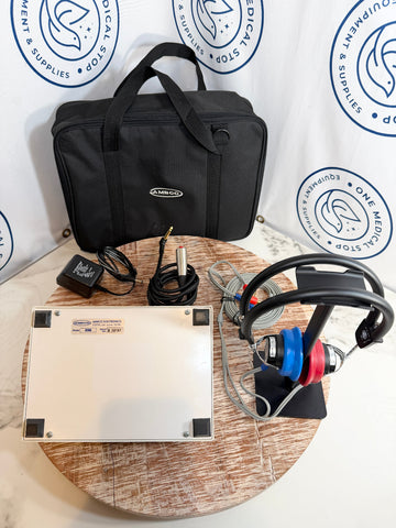 AMBCO audiometer with matching black carrying case, shown with accessories including a power adapter, cables, and over-ear headphones on a round wooden table in front of a branded white backdrop.