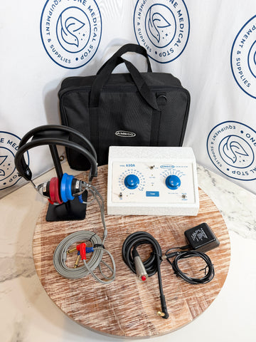 AMBCO audiometer kit displayed on a round wooden tray, including a white control unit with blue dials, a black headphone on a stand, coiled gray and black cables, a black carrying case, and a black power adapter. 